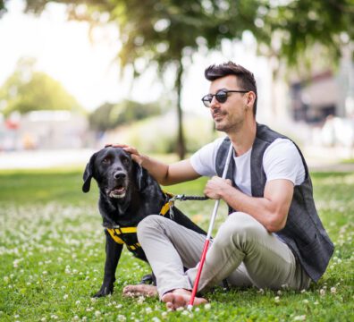Young blind man with white cane and guide dog sitting in park in city, resting.