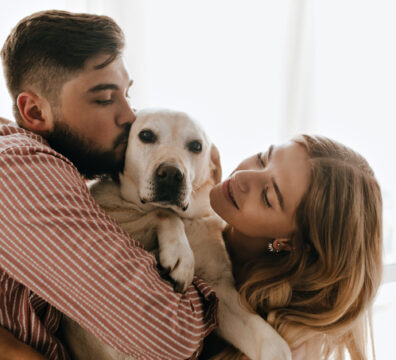 Romantic couple in good mood plays and hugs white dog. Man kisses his labrador against window